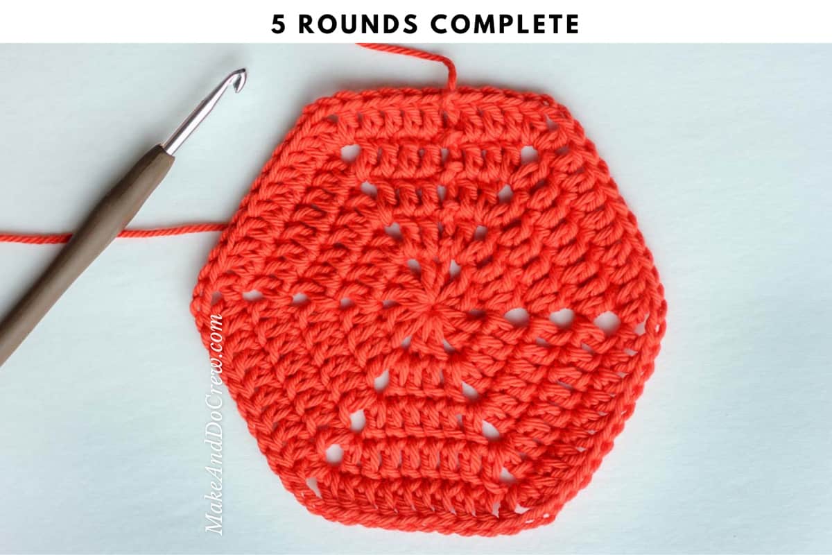 A brown crochet hook next to a completed crochet hexagon made with orange yarn.