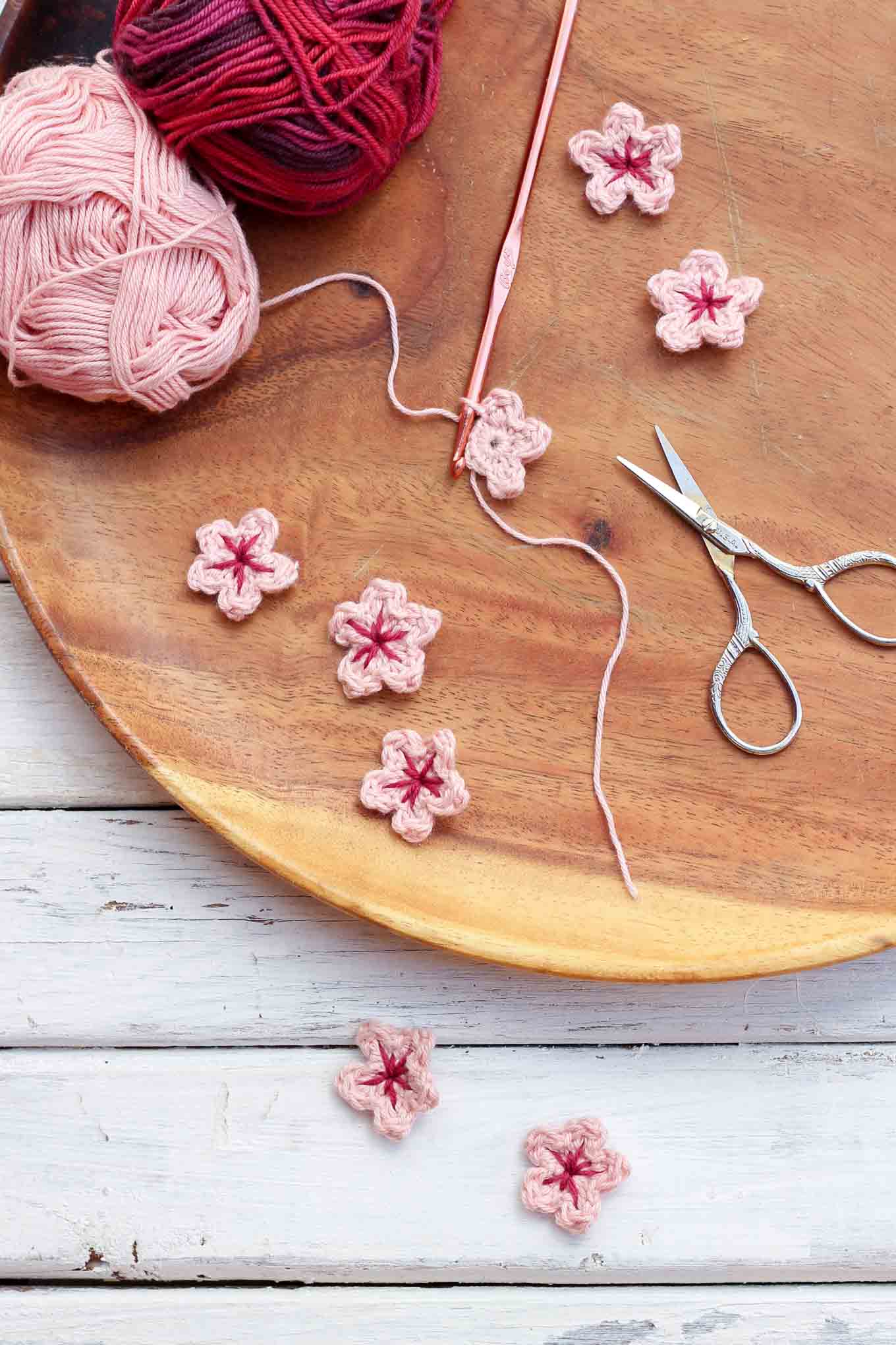 Mini crochet cherry blossoms on a wooden plate with yarn skeins, hook and scissors.