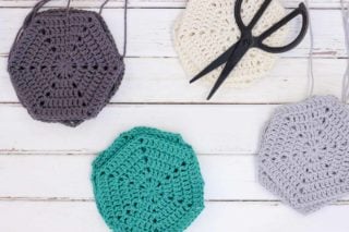 Overhead view of stacks of crochet hexagons in the colors grey, teal, cream and light grey. A utilitarian pair of scissors sits atop on stack of hexagons.