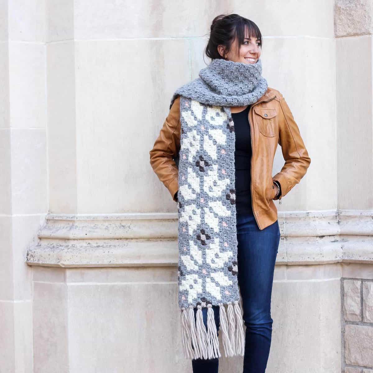A woman looking sideways smiling with her hands on a brown leather jacket and a gray Nordic crochet super scarf on her neck.