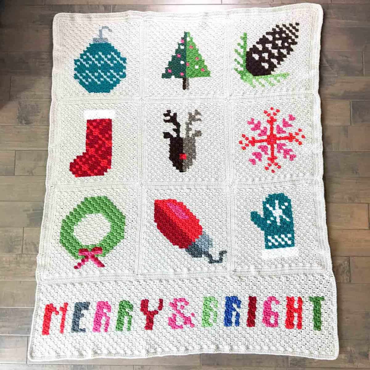 Flat lay of a white and Christmas-themed C2C blanket with the words merry & bright at the bottom is placed on a wooden floor.