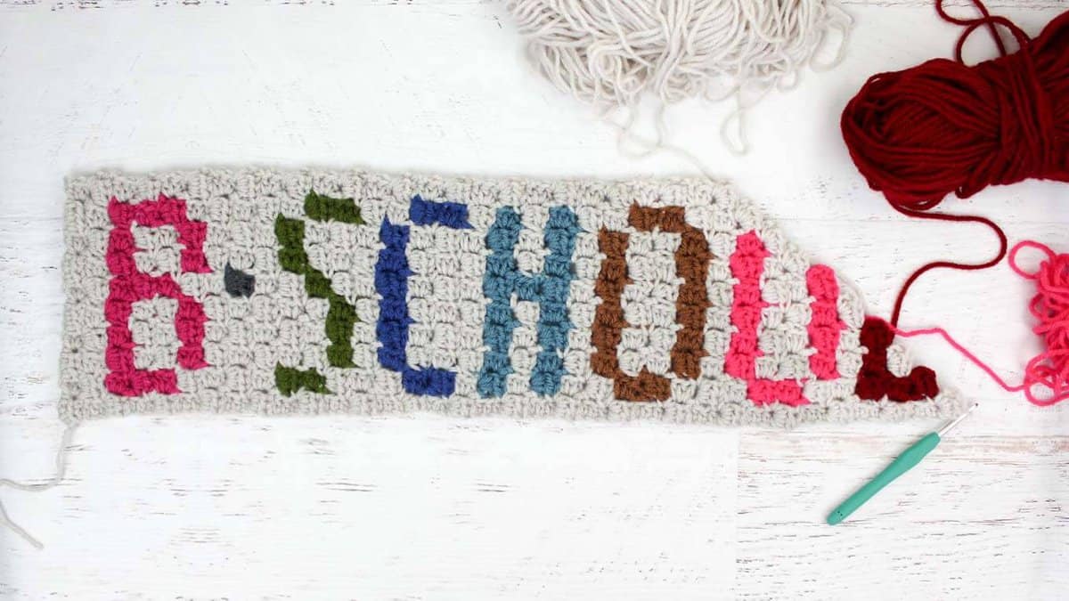 Flat lay of a C2C blanket with colorful letters on it with white, red, and pink yarn and a crochet hook on a white table.