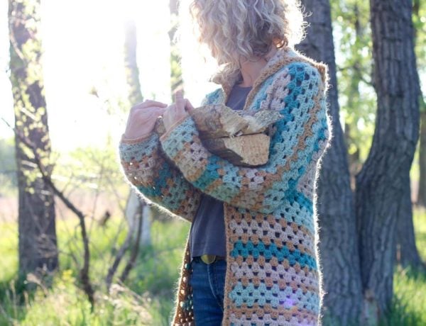 Blonde woman carrying firewood in the woods, wearing a crochet cardigan made from hexagons.