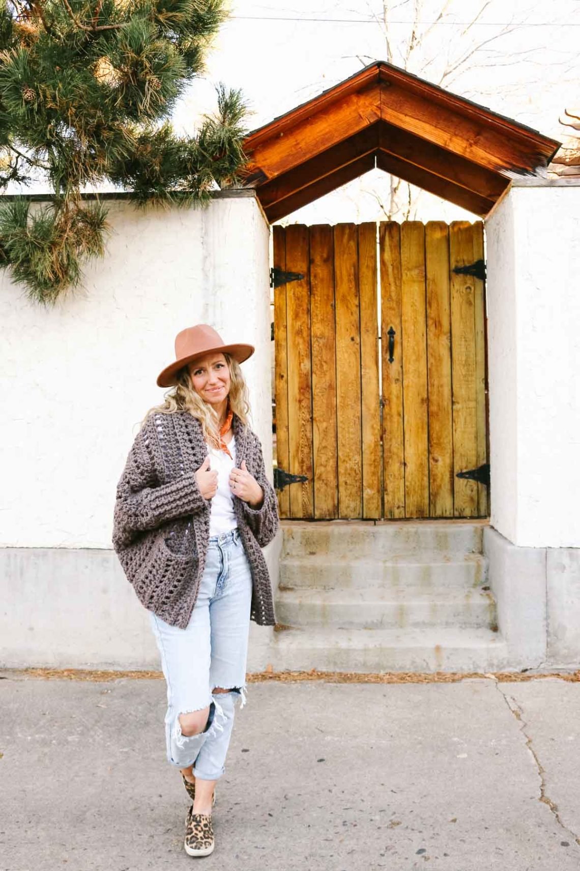 This image shows a blonde woman standing in front of a gate and a white wall. She is wearing a brownish grayish crochet cardigan with pockets, light blue jeans, a white shirt and a tan hat. Her hands are in her pockets and she is looking at the camera.