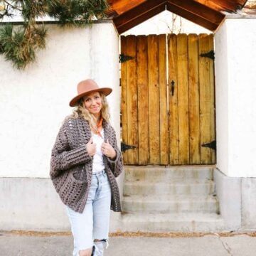This image shows a blonde woman standing in front of a gate and a white wall. She is wearing a brownish grayish crochet cardigan with pockets, light blue jeans, a white shirt and a tan hat. Her hands are in her pockets and she is looking at the camera.