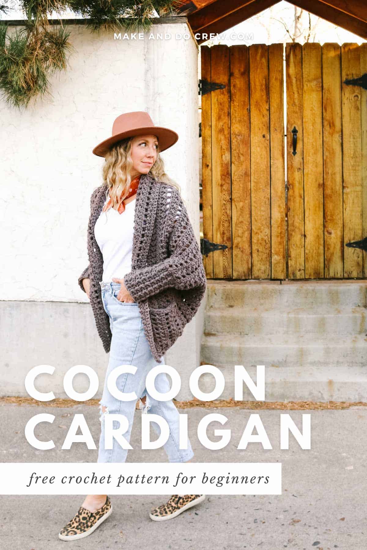 This image shows a blonde woman standing in front of a gate and a white wall. She is wearing a brownish grayish crochet cardigan with pockets, light blue jeans, a white shirt and a tan hat. Her hands are in her pockets and she is looking over her left shoulder.