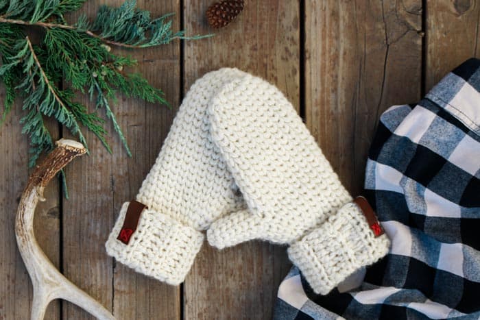 A pair of chunky mittens on a table with a button down plaid, a tree branch and a pinecone.
