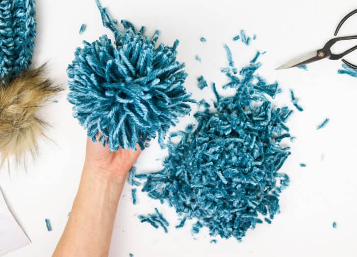An overhead shot of a hand holding the finished pom poms with excess, a finished hat, and scissors on the white background. 