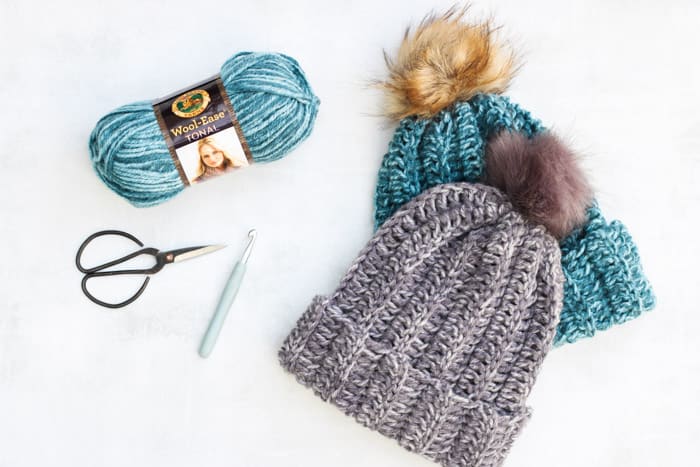Flat lay with two crochet beanies teal and gray colored, yarn, crochet hook, and scissors on a white background.