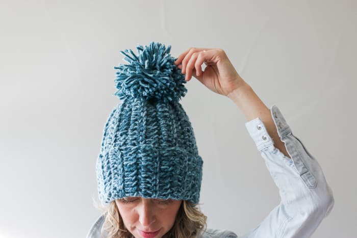 A woman wearing the finished green crochet hat with her hand on the pom poms with a white background.