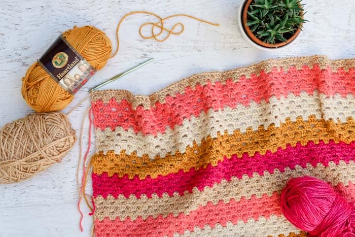 An overhead view of a crochet granny stripe blanket made in pink, magenta, mustard and cream colored Lion Brand Heartland yarn.