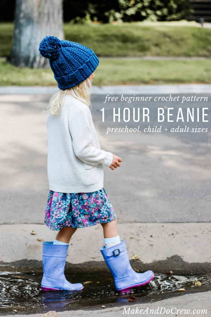 A kid wearing a crochet hat and a pair or rain boots standing on a puddle of water.