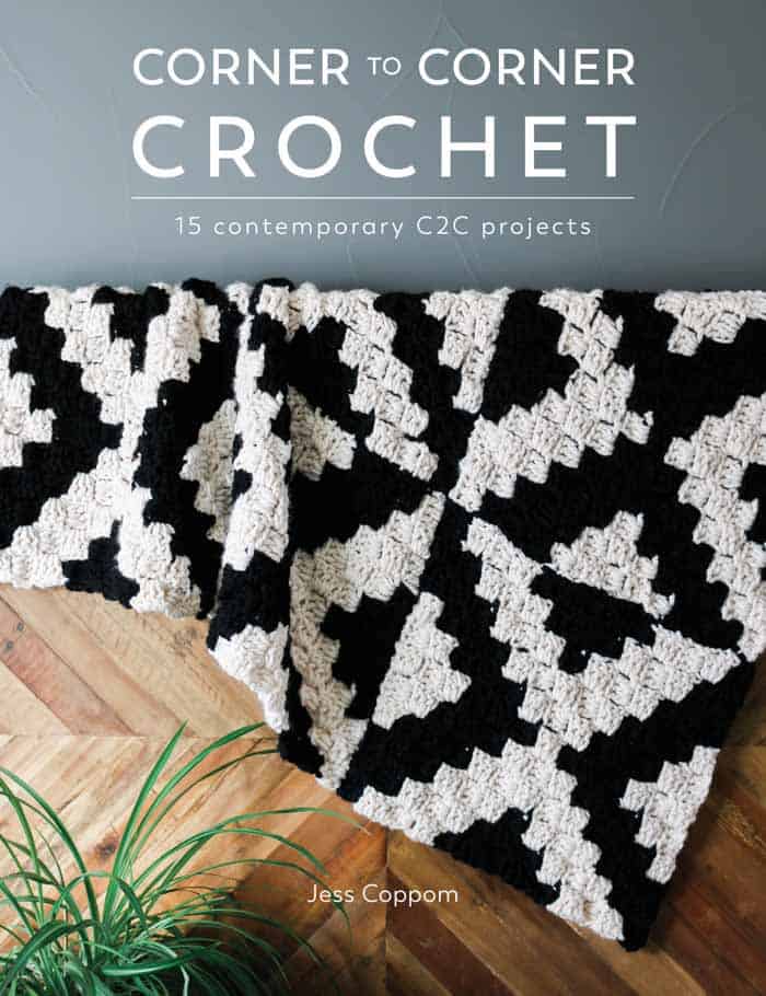 A black and white corner-to-corner crochet blanket draped on a wooden table with a plant at the bottom and a gray wall.