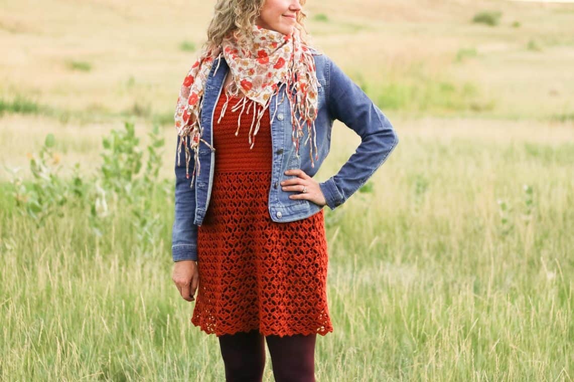 Woman standing in a field wearing a crochet dress, patterned scarf and jean jacket.