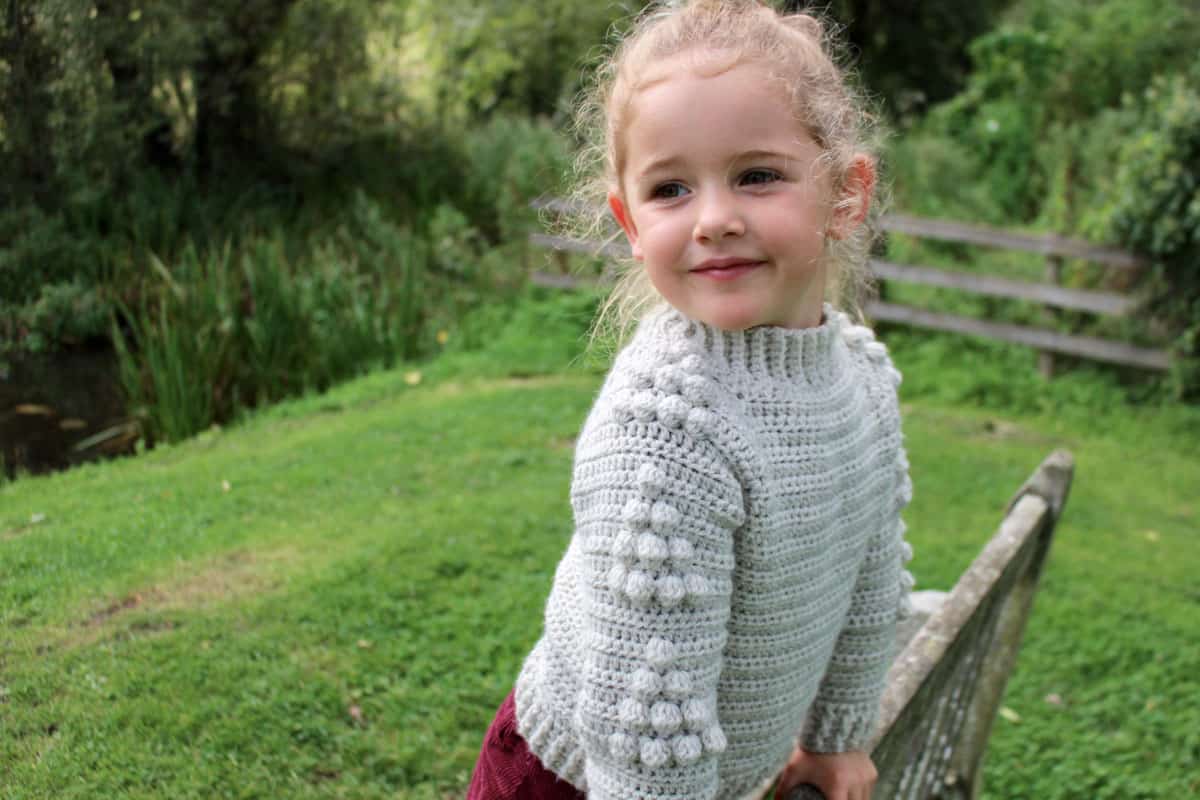 Young girl in green grass leaning on a bench. She is wearing a crochet pullover sweater, and the sleeves show a tree bobble motif. Free pattern and tutorial.