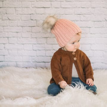 An adorable baby girl wearing a crochet hat with a fur pom pom.