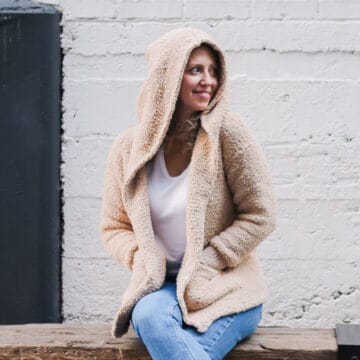 Woman sitting on a bench wearing a hooded crochet jacket made from Lion Brand Baby Soft Boucle yarn.