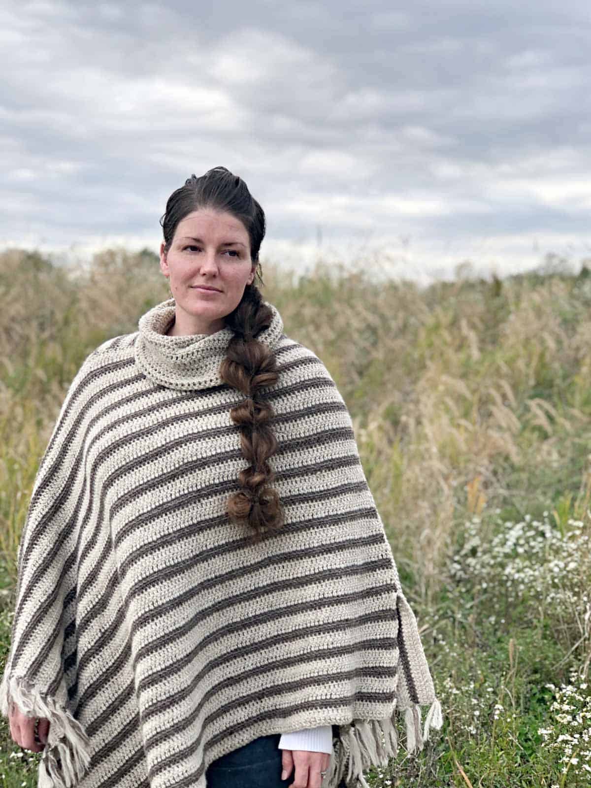Women standing in field. She is wearing a striped, asymmetrical crochet poncho with fringe and a dark denim skirt.