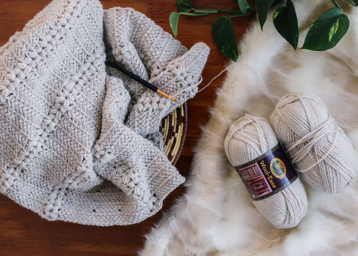 Off white crochet sweater made with herringbone double crochet and puff stitches laying in a basket next to balls of Lion Brand Wool-Ease.
