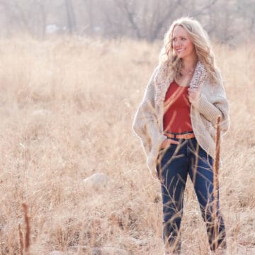 Woman standing in a field of natural grasses wearing a drapey, dolman-sleeved crochet cardigan with faux fur trim.