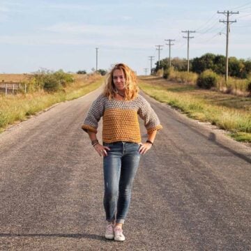 Girl standing in road surrounded by fields, wearing a cropped crochet sweater with ¾ length sleeves.