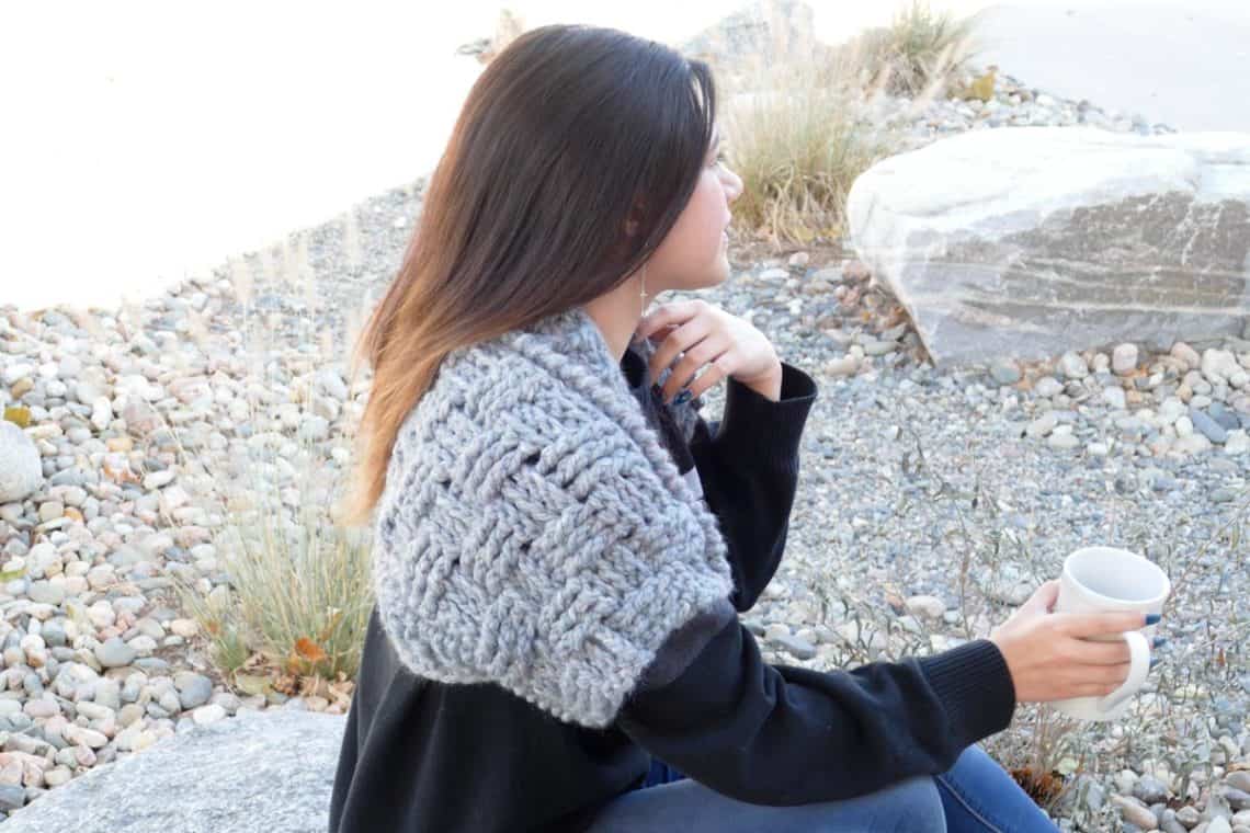 Women with long dark hair sitting on a rock, facing away from the camera and holding a white mug. She is wearing blue jeans, a long-sleeved blank shirt, and a chunky gray crochet bolero shrug.