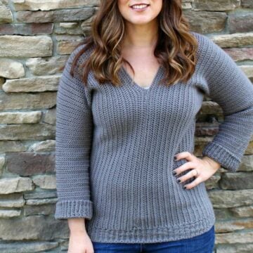 Girl with long brown hair, standing in front of brick wall, wearing a gray crochet v-neck sweater.
