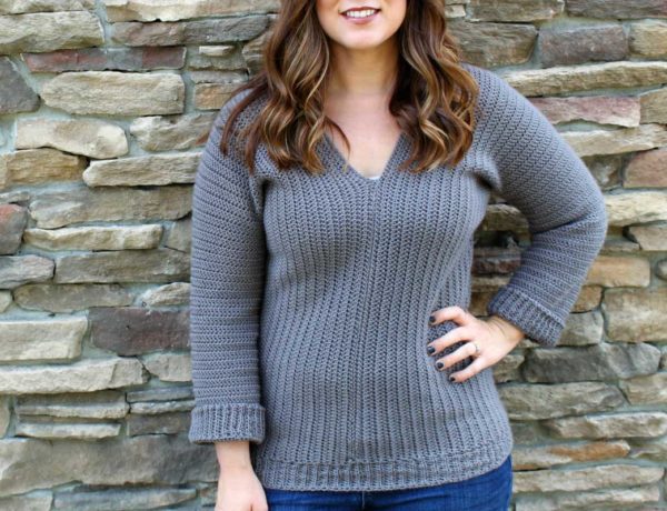 Girl with long brown hair, standing in front of brick wall, wearing a gray crochet v-neck sweater.