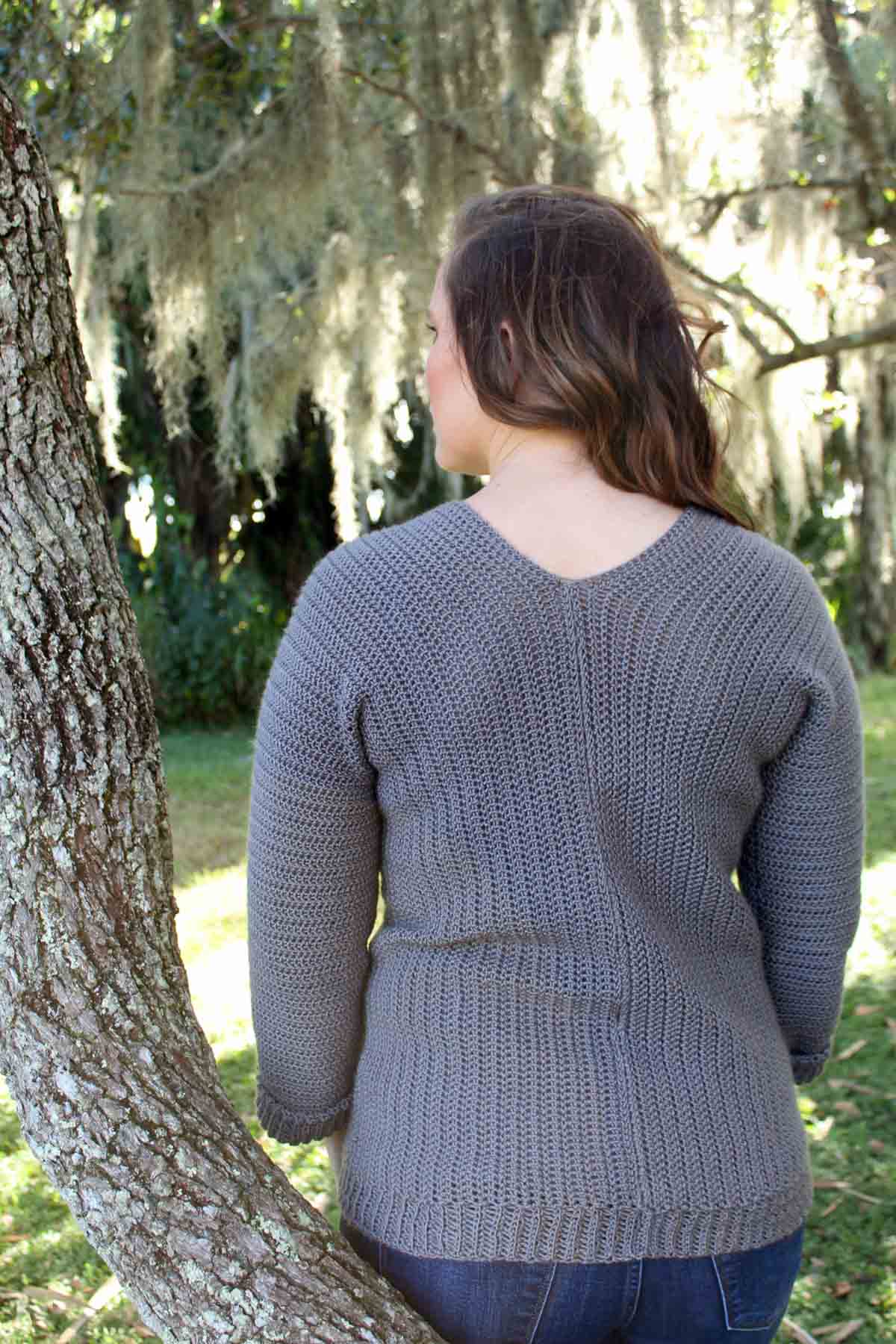 Woman standing outside next to tree. She is wearing a gray v-neck crochet pullover sweater. Free pattern for the sweater.