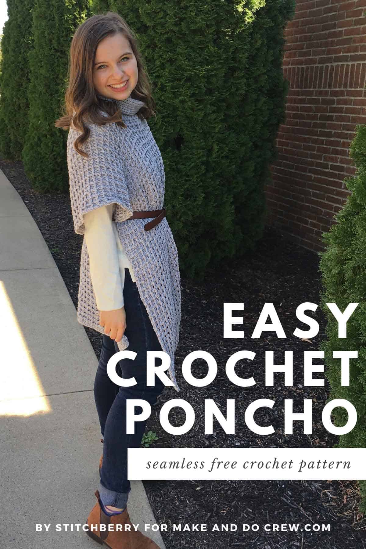 Woman standing on sidewalk on a sunny day in front of a brick wall and tree. She is wearing jeans, a long sleeve white shit, and a long gray crochet poncho with a brown belt cinched around her waist.