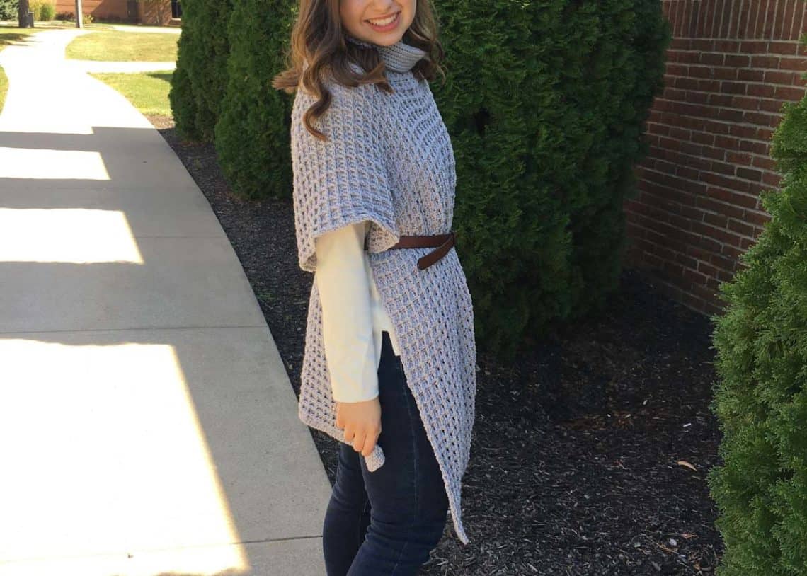 Woman with medium length brown hair standing on sidewalk on a sunny day in front of a brick wall and tree. She is wearing jeans, a long sleeve white shit, and a long gray crochet poncho with a cowl neck. She has a brown belt cinched around her waist.