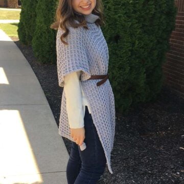 Woman with medium length brown hair standing on sidewalk on a sunny day in front of a brick wall and tree. She is wearing jeans, a long sleeve white shit, and a long gray crochet poncho with a cowl neck. She has a brown belt cinched around her waist.