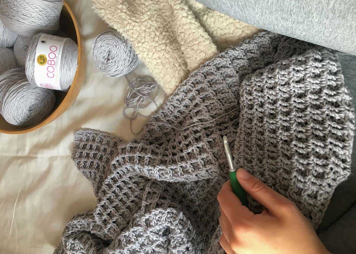 This photo shows an in-progress shot of a gray crochet poncho made using the waffle stitch. There is a bowl of yarn filled with Lion Brand Coboo yarn in the Silver coloway. 