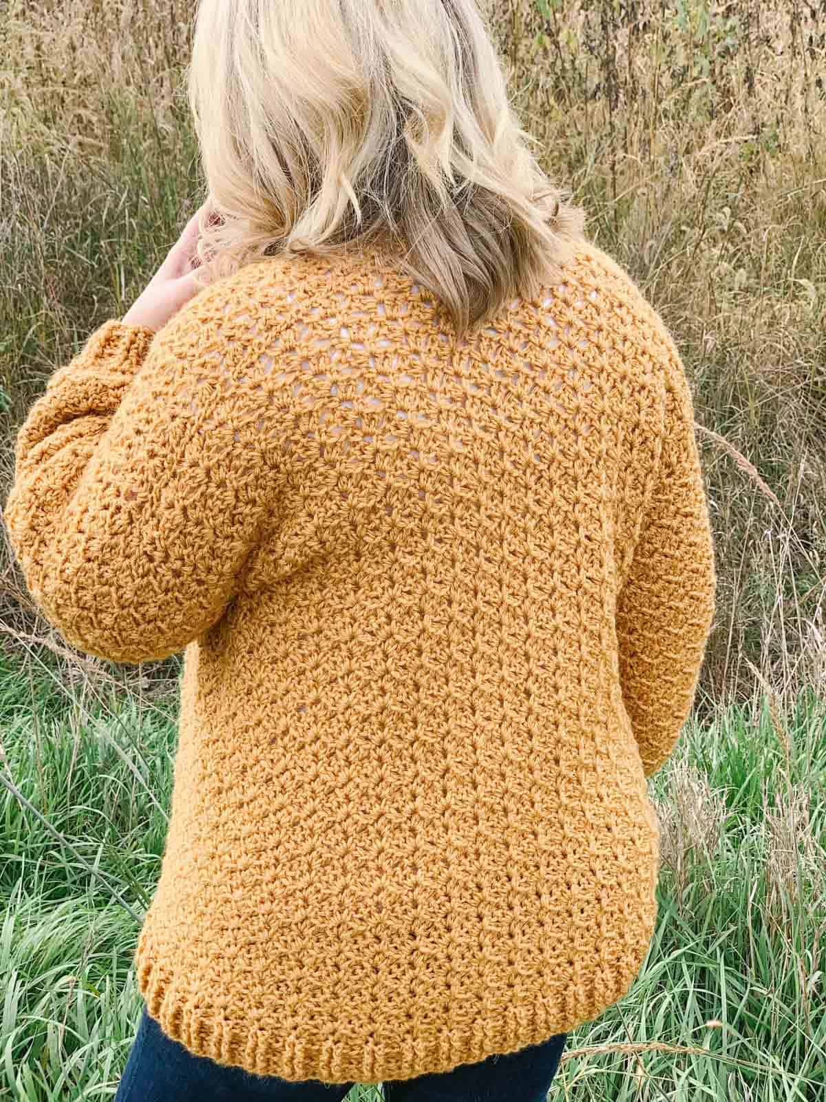 Woman with blonde hair standing in field with a mustard yellow crochet cardigan on.