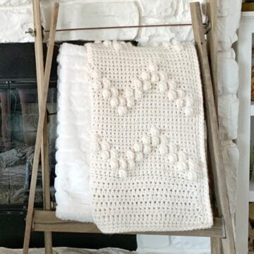 Cream colored crochet bobble blanket displayed on a blanket ladder in front of a fireplace.