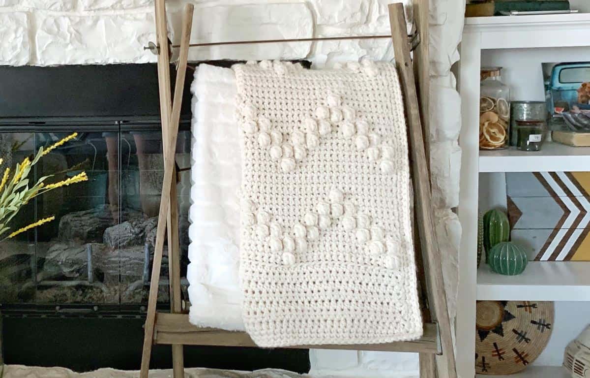 Cream colored crochet bobble blanket displayed on a blanket ladder in front of a fireplace.