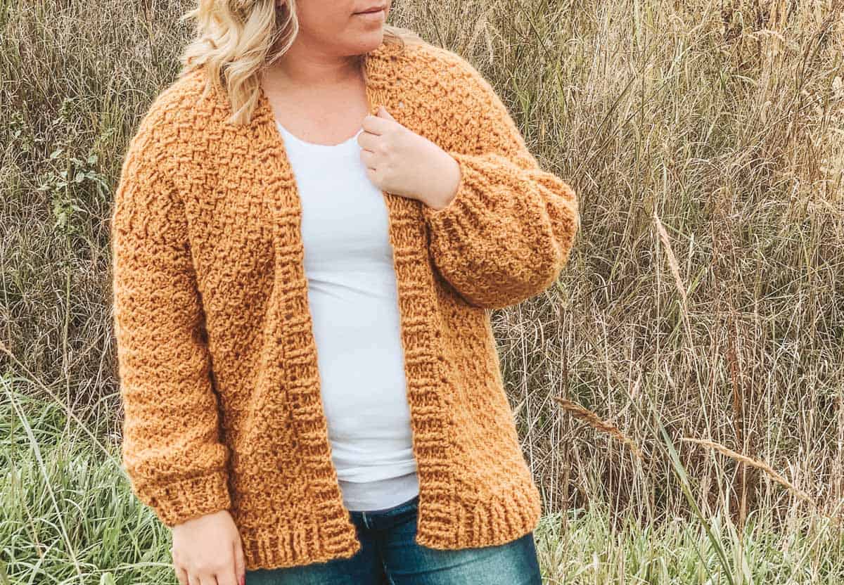 Woman with blonde hair standing in field of grass. She is wearing blue jeans, a white shirt and a mustard yellow crochet cardigan.