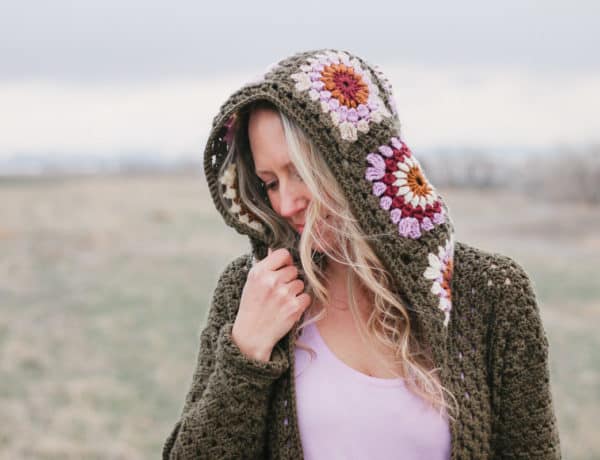 A blonde woman wearing a hooded crochet sweater featuring retro crochet granny square motifs in modern colors of Lion Brand Basic Stitch yarn.