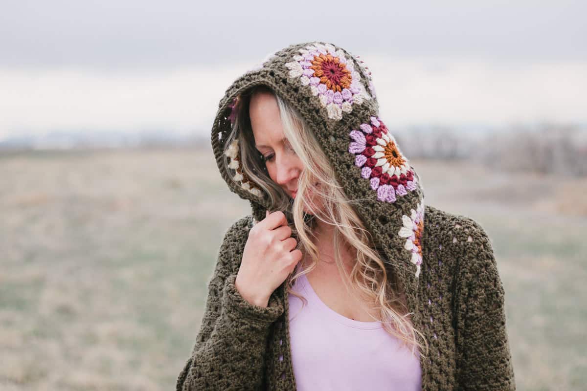 A woman wearing and holding the hood of a retro crochet granny square cardigan.