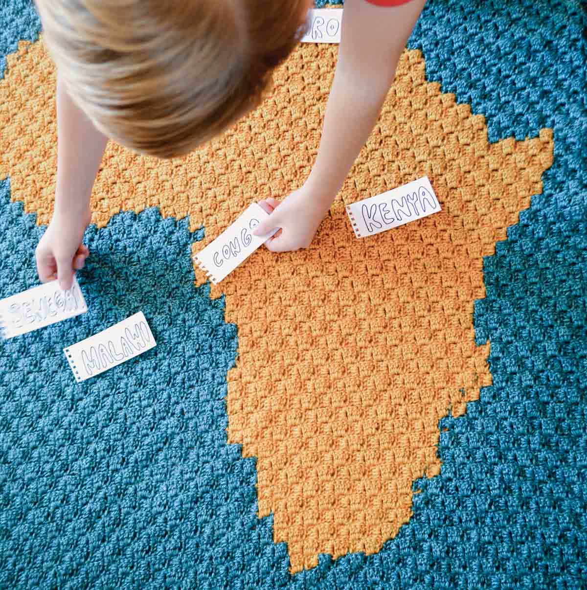 An overhead shot of a child placing labels of cities on a crochet African map blanket.