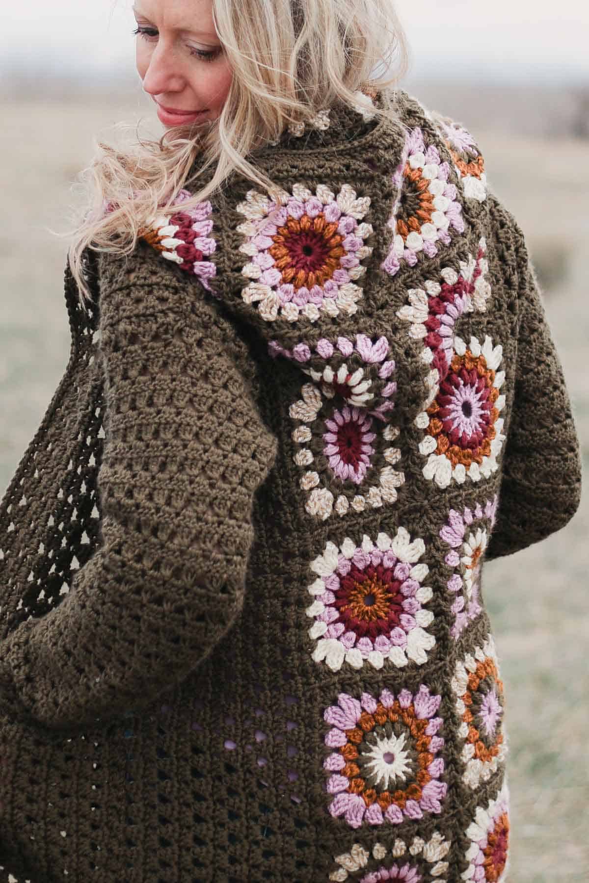 A woman showing the back of a vintage-looking crochet granny square cardigan with a hood and pockets.