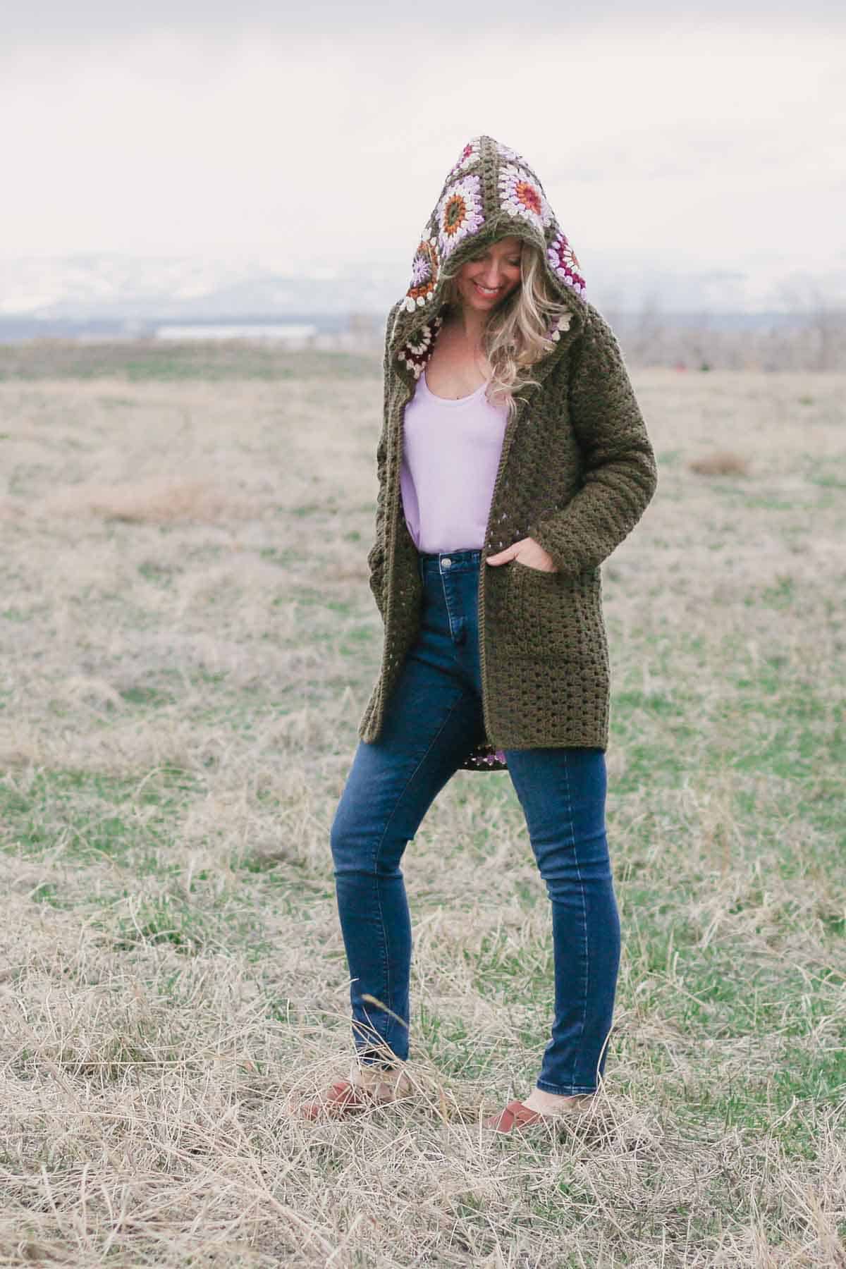 A woman looking down and smiling with her hands in the pockets of a modern crochet granny square cardigan with a hood.