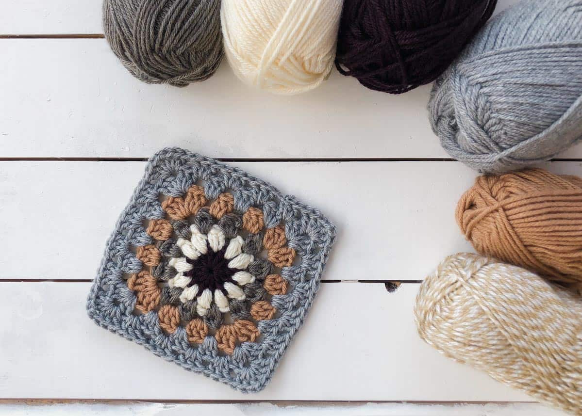 Flat lay of a crochet square pattern with six neutral-colored anti-pilling yarns on top of a white table.