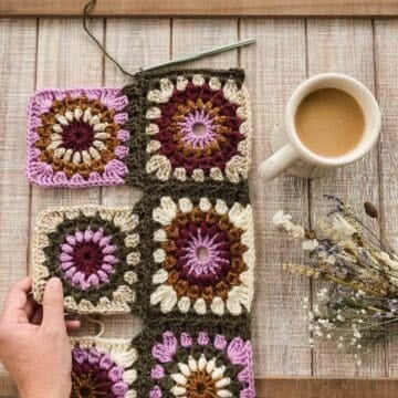 An overhead view of purple, green and cream colored sunburst granny squares in the progress of being joined with crochet stitches. A woman's hand is reaching into frame and a cup of coffee sits nearby.