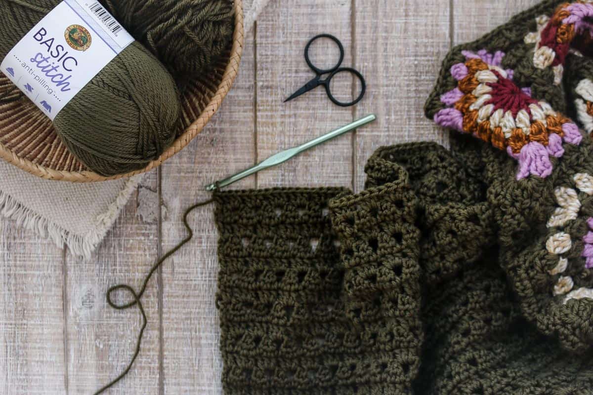 Flat lay of an olive green cardigan with a metal crochet hook, black scissors, and a wooden basket with an anti-pilling yarn.