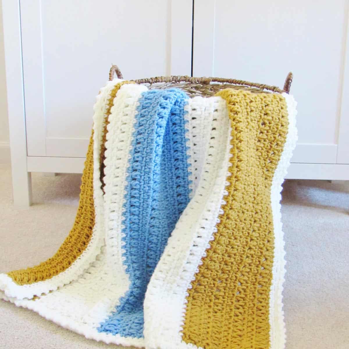 A wide gold, light blue, and white colored one-day crochet blanket pattern for beginners inside a wicker basket on the floor.