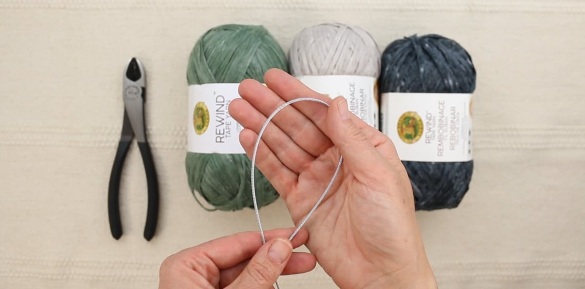 A hand holding galvanized wire rope above three skeins of Lion Brand Rewind yarn.