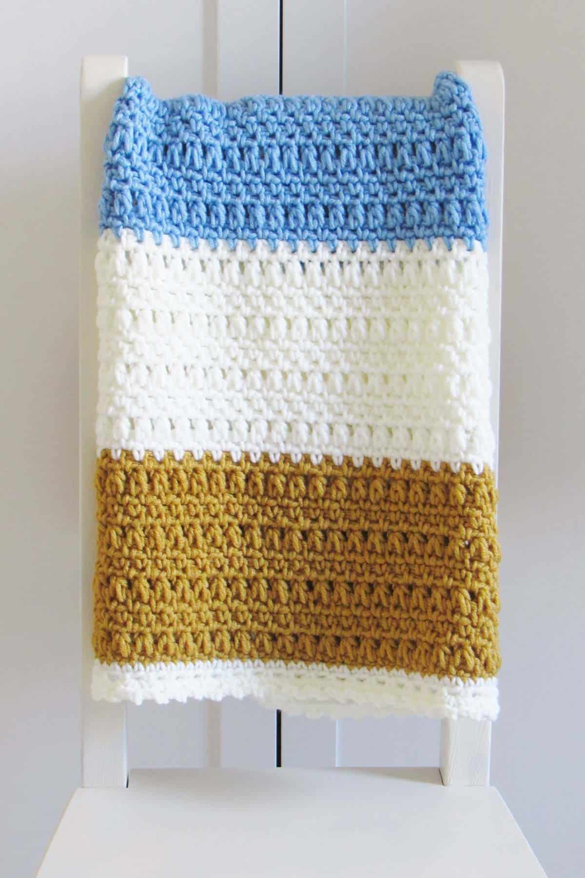 A wide gold, light blue and white colored one day crochet blanket pattern for beginners folded in a white chair.