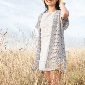 Women standing in field wearing a white dress and a lacy gray crochet cardigan. Her hand is touch the back of her sun hat.