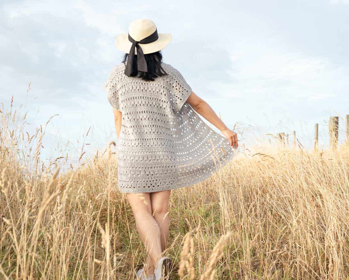 Women in a field facing away from the camera. She is wearing a tan and black sun hat, a white dress and a gray crochet cardigan. Her hand is holding one side of the cardigan spread out.